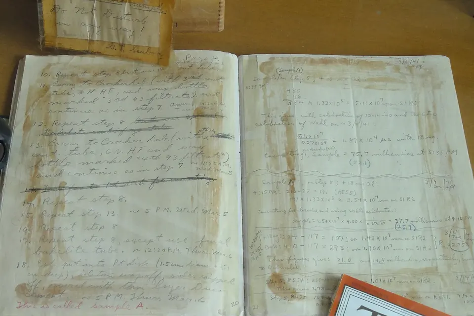 An old bound laboratory logbook lying open on a desk, its handwritten pages yellowed and stained