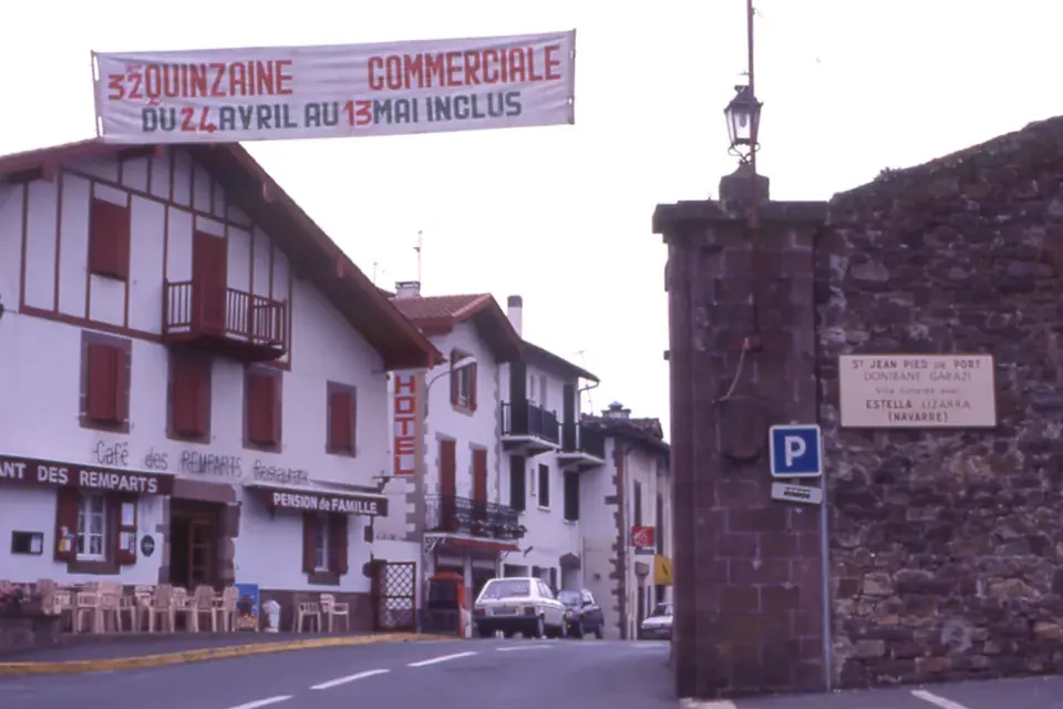 A street at the entrance of a small town with half-timbered houses, a banner over the road and a stone gate bearing a bilingual sign
