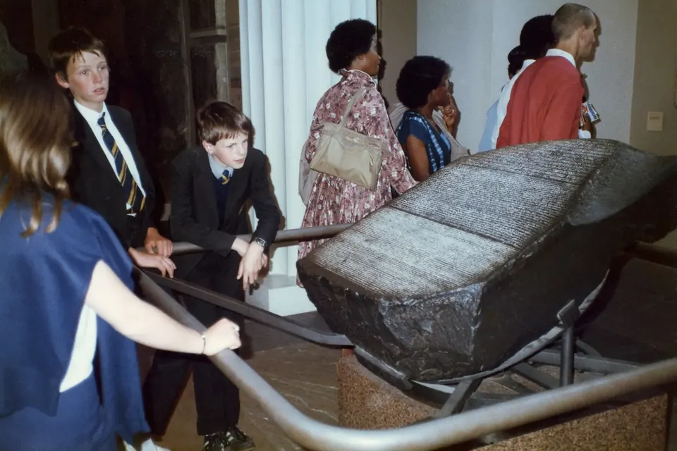 Museum visitors leaning on a rail to look at the Rosetta Stone, a dark inscribed slab displayed at an angle