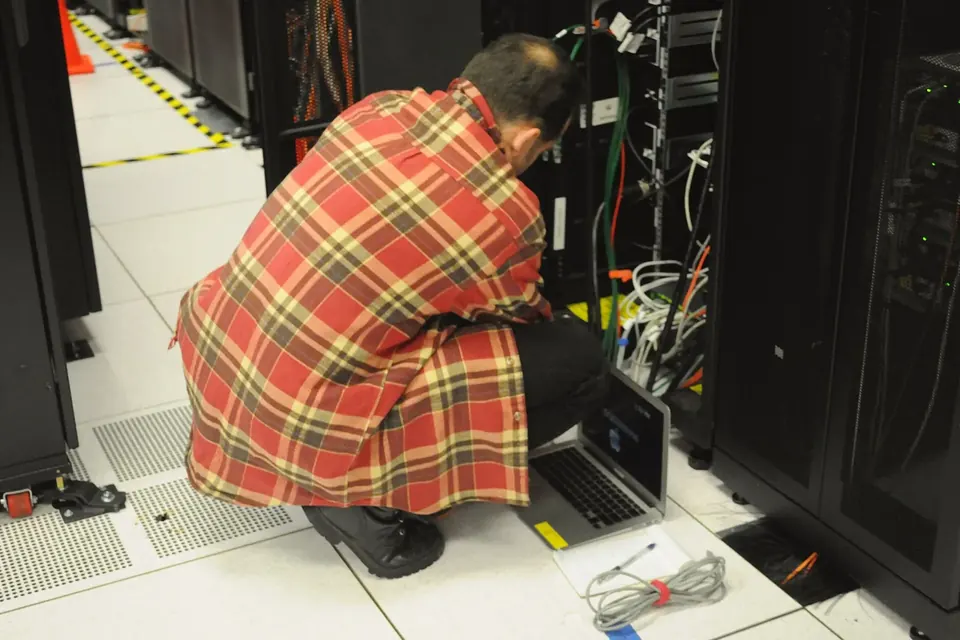 A technician in a checked shirt crouches with a laptop in front of an open server rack in a data centre