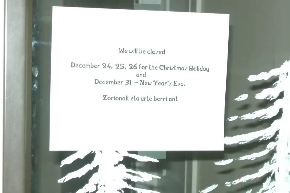 A printed notice taped to a glass door announcing holiday closing dates in English, with a greeting in Basque below