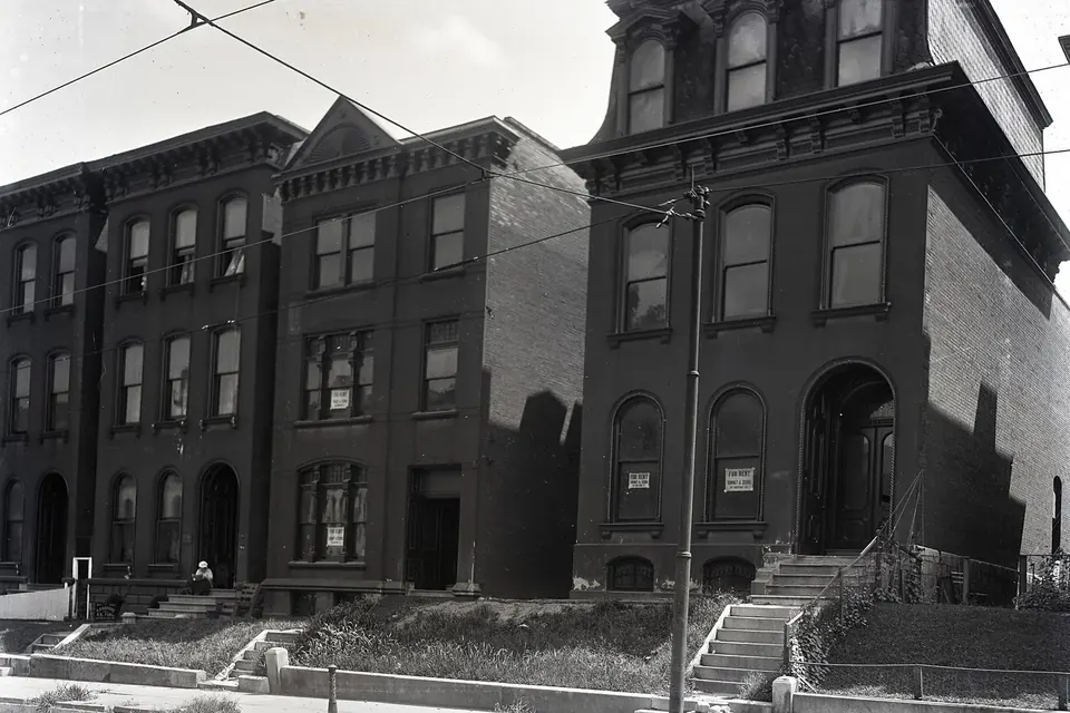 A row of three-storey brick apartment houses with bay windows and small rental signs in the windows, in an old black-and-white photograph